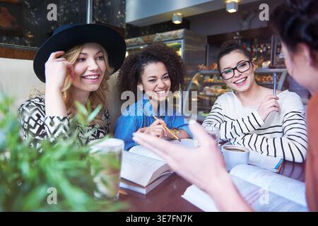 Lächelnde Gruppe von Freundinnen, die miteinander reden, während sie im Café mit Büchern am Tisch sitzen Stockfoto