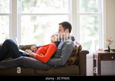 Frau mit gemischter Rasse, die einen Laptop benutzt, während sie zu Hause auf der Couch ihres Freundes liegt Stockfoto