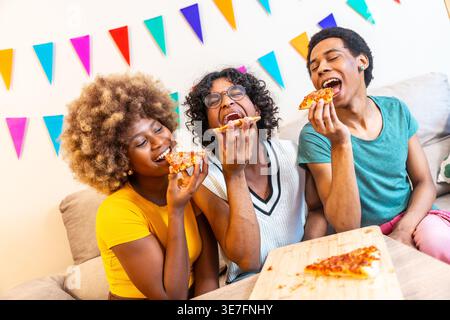Fröhliche, vielfältige Gruppe von Freunden, die es genossen, ein Stück Pizza zu beißen, während sie zu Hause auf dem Sofa sitzen Stockfoto