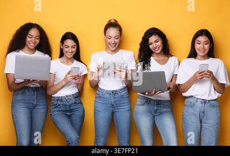 Verschiedene Frauengruppen, die Hemden und Jeans mit verschiedenen elektronischen Geräten tragen und im Studio vor gelbem Hintergrund stehen Stockfoto