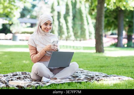 Lächelnde indische muslimische Frau im Hijab mit Smartphone, die tagsüber mit Laptop auf dem Schoß auf einer karierten Decke im Park sitzt Stockfoto