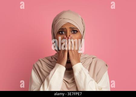 Asiatische Frau im Hijab, die den Mund mit Händen bedeckt, während sie im Studio vor rosa Hintergrund steht Stockfoto