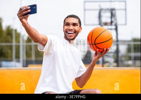 Lächelnder Schwarzer Mann, der tagsüber auf Beton sitzend im Basketballfeld im Park Selfie macht Stockfoto