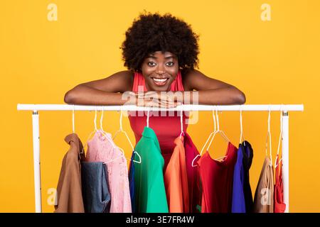 Schwarze Frau mit Afro, die sich auf Kleiderständer stützt, während sie im Studio vor gelbem Hintergrund steht Stockfoto