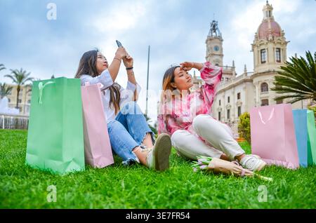Asiatische Frau, die tagsüber mit dem Smartphone Fotos macht, während sie mit einer Freundin auf dem Gras sitzt, umgeben von bunten Einkaufstaschen im Stadtpark... Stockfoto