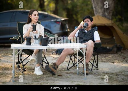 Asiatisches Paar, das beim Camping im Wald an einem Klapptisch in der Nähe des Zeltes sitzt Stockfoto