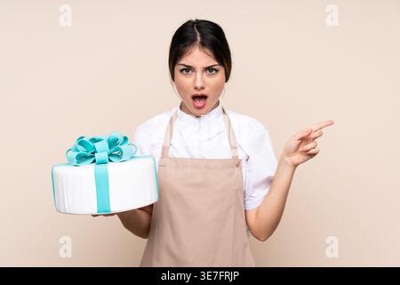 Schockierte hispanische Konditorin, die mit dem Finger zur Seite zeigt und Kuchen im Studio vor beigem Hintergrund hält Stockfoto