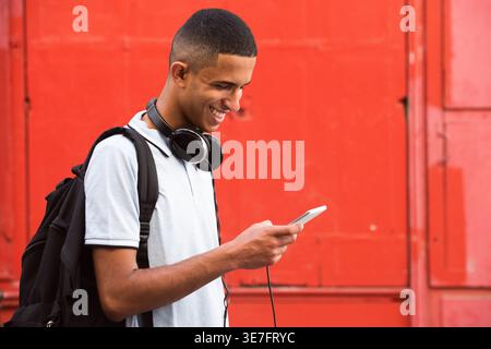 Lächelnder arabischer männlicher Teenager mit Kopfhörern am Hals mit Smartphone, das an der roten Wand steht Stockfoto