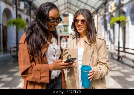 Schwarze und kaukasische Freundinnen tragen Trenchcoat und Sonnenbrille und überprüfen das Smartphone, während sie tagsüber in der Stadt stehen Stockfoto