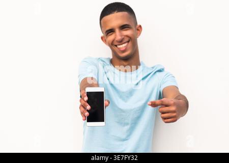 Lächelnder arabischer männlicher Teenager, der mit dem Finger auf das Smartphone zeigt, das vor weißem Hintergrund steht Stockfoto