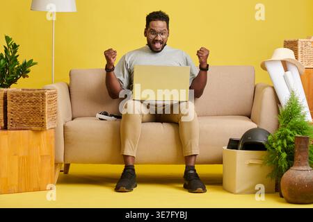Schockierter asiatischer Mann mit Brille und offenem Mund, der geschlossene Fäuste hochhebt und auf den Laptop blickt, der auf dem Sofa zu Hause sitzt Stockfoto