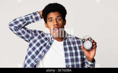 Schockierter schwarzer Mann mit Hand hinter Kopf, der den Wecker hält, der im Studio vor weißem Hintergrund steht Stockfoto