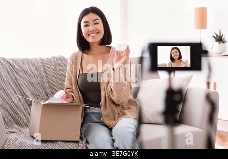 Asiatische Frau, die ein Video zur Aufnahme von Schönheitsprodukten für ihren Beauty-Blog hält und zu Hause auf dem Sofa sitzt Stockfoto