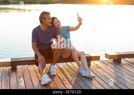 Ein lächelndes chinesisches Paar mittleren Alters macht Selfie, während es bei Sonnenuntergang auf einer Betonbank in der Nähe des Sees im Park sitzt Stockfoto