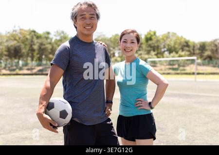 Ein lächelndes chinesisches Paar mittleren Alters hält Fußball, während es an sonnigen Tagen auf dem Fußballfeld steht Stockfoto