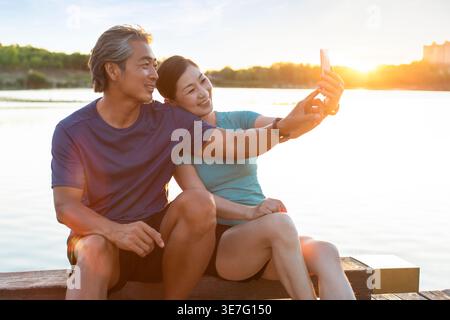 Ein lächelndes chinesisches Paar mittleren Alters macht Selfie, während es bei Sonnenuntergang auf einer Betonbank in der Nähe des Sees im Park sitzt Stockfoto