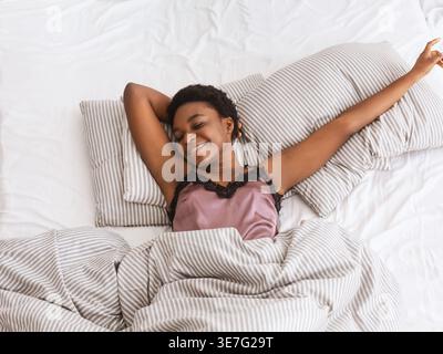 Faulheit und guten Morgen, schlafen Sie am Sonntagmorgen und genießen Sie den Urlaub. Glückliche junge afroamerikanerin liegt auf dem Bett und dehnt sich auf weichen Kissen, wh Stockfoto