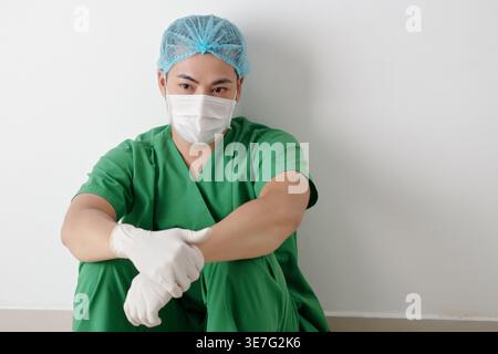 Müde asiatische männliche medizinische Fachkraft in Schutzhandschuhen, die kurze Pause beim Sitzen und in die Ferne im Krankenhaus blicken Stockfoto