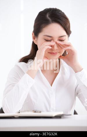 Gestresste chinesische Geschäftsfrau mit Händen unter den Augen und geschlossenen Augen, die am Tisch im Büro sitzt Stockfoto
