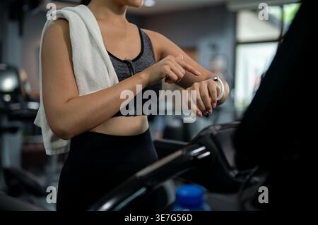 Thailändische Frau in Fitnesskleidung, die ihre Smartwatch beim Gehen auf dem Laufband im Fitnessstudio festlegt Stockfoto