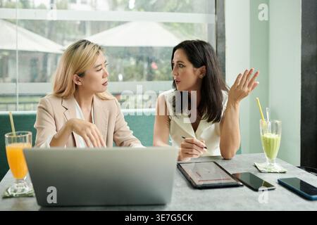 Ernsthafte asiatische und weiße Geschäftsfrauen diskutieren während der Pausen im Café über Arbeitsdetails Stockfoto