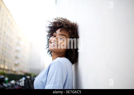 Lächelnde schwarze Frau, die über die Schulter schaut und sich an die weiße Wand auf der Stadtstraße lehnt Stockfoto