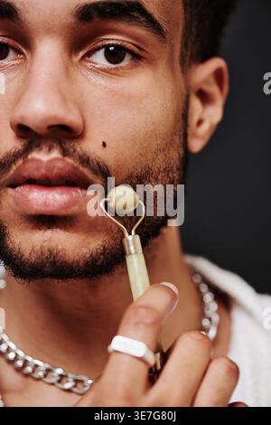 Gemischter bärtiger Mann mit Jadewalze auf Gesicht, der vor schwarzem Hintergrund im Studio steht Stockfoto