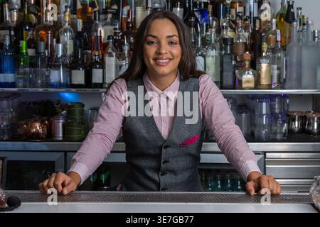 Lächelnde hispanische Barkeeperin, die in der Bar stand und sich die Hände auf die Theke lehnte Stockfoto