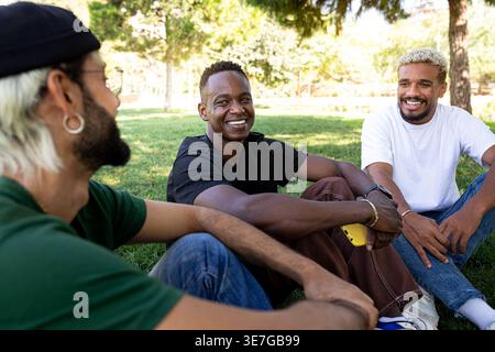 Fröhlicher Schwarzer Mann, der ein Smartphone hält und mit hispanischen und kaukasischen männlichen Freunden plaudert und lacht, die am sonnigen Tag im Park auf Gras sitzen Stockfoto