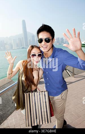 Ein lächelndes chinesisches Paar mit Sonnenbrille und winkenden Händen, während es tagsüber am Hafen steht Stockfoto
