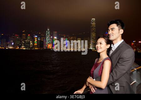 Ein lächelndes chinesisches Paar umarmt sich und blickt weg, während es nachts am Hafen steht Stockfoto