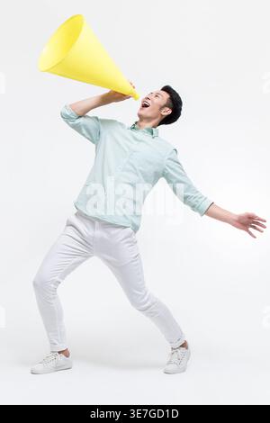 Chinesischer Mann, der nach Megaphon schreit und wegschaut, während er vor weißem Hintergrund im Studio steht Stockfoto