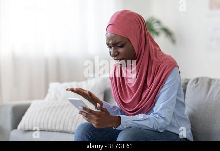 Verwirrte Schwarze Frau, die Hijab mit Smartphone trägt, während sie zu Hause auf dem Sofa sitzt Stockfoto