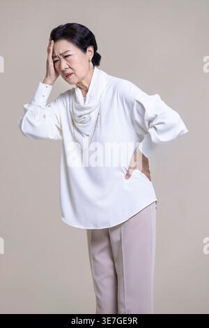 Gestresste chinesische Seniorin, die wegschaut und Kopfschmerzen hat, die mit Händen auf Stirn und Taille vor braunem Hintergrund im Studio stehen Stockfoto