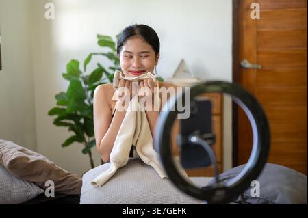 Lächelnde asiatische Frau nimmt sich auf, ihre Kleidung vor der Kamera zu zeigen und zu sehen, während sie zu Hause auf dem Sofa sitzt Stockfoto