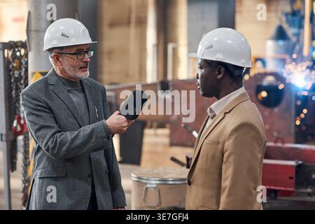 Schwarzer männlicher Ingenieur, der einen Sicherheitshelm trägt und einen kaukasier befragt, der im Lager steht Stockfoto