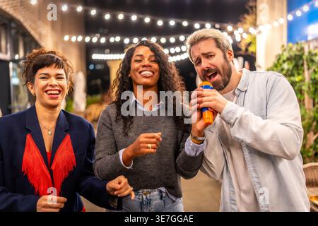 Verschiedene Freunde halten ein Mikrofon und singen Karaoke zusammen, während sie abends auf der Dachbar stehen Stockfoto
