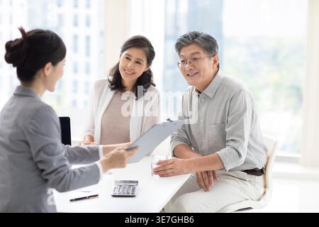 Lächelndes chinesisches Paar, das mit einer chinesischen Finanzberaterin spricht, während er am Tisch im Büro sitzt Stockfoto