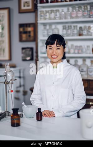 Japanische Apothekerin, die mit Händen auf der Arbeitsheke steht, mit Mörtel, Stößel, Trenntrichter und Flaschen mit Chemikalien in Vintage... Stockfoto