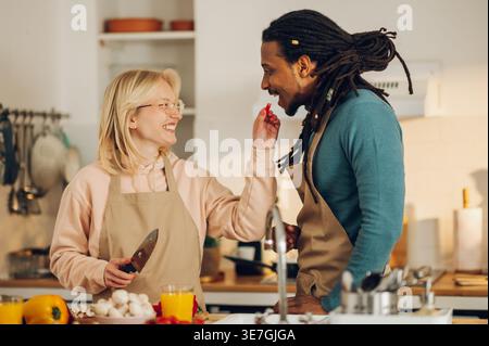 Lächelnder Mixed Race Mann mit Schlössern und kaukasische Freundin füttert ihm Tomaten zusammen am Tisch mit Gemüse in der Küche zu Hause Stockfoto