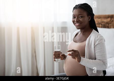Schwarze schwangere Frau mit Vitaminpille Kapsel und Glas Wasser, während sie zu Hause auf dem Bett im Schlafzimmer sitzt Stockfoto