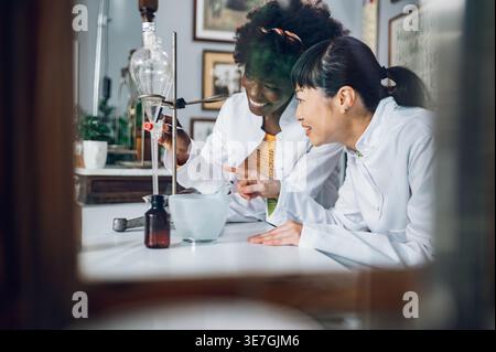 Schwarze Chemikerinnen öffnen das Ventil des Trenntrichters mit einer japanischen Kollegin, die die Flüssigkeit beobachtet und auf den alten ph-wert zeigt... Stockfoto