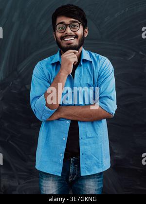 Lächelnder indischer Mann in blauen Langarmen mit Brille, die mit Hand am Kinn an der Tafel im Klassenzimmer steht Stockfoto