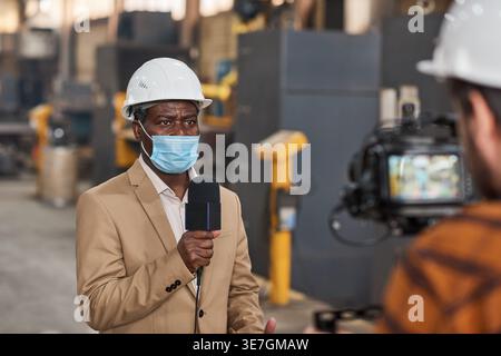 Schwarzer Journalist mit Schutzhelm und Gesichtsmaske mit Mikrofon im Lager Stockfoto