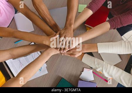 Verschiedene Studentengruppen stapeln die Hände in kreisförmiger Form zusammen, während sie am Tisch in der Bibliothek sitzen Stockfoto