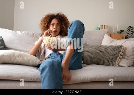Hispanische Frau mit lockigem Haar isst Popcorn und schaut fern, während sie zu Hause auf dem Sofa sitzt Stockfoto