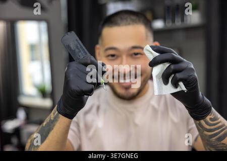 Lächelnder asiatischer Tätowierer, der in seinem Tätowierstudio eine Schreibmaschine und ein Taschentuch hält Stockfoto