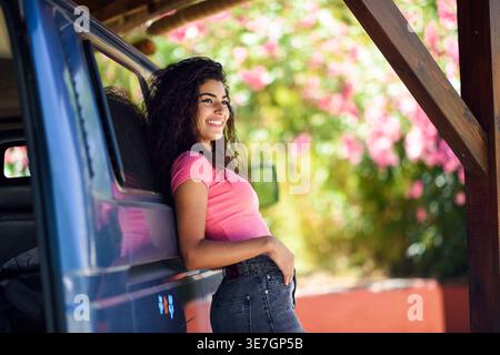Lächelnde arabische Frau mit lockigen Haaren in rosafarbenem Hemd und Jeans, die wegblickt, während sie sich an einen Wohnwagen lehnt, der in der Garage geparkt ist Stockfoto
