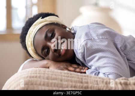 Lächelnde hispanische Frau mit Afro-Stirnband, die zu Hause auf dem Bett liegt Stockfoto