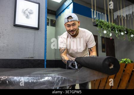 Lächelnder asiatischer Tätowierer mit Mütze und Latexhandschuhen, der die Couch für die Tätowierung im Studio vorbereitet Stockfoto